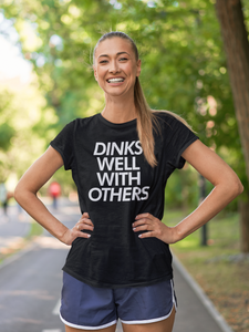smiling woman in athletic shorts at park wearing a black dinks well with others pickleball shirt apparel front view